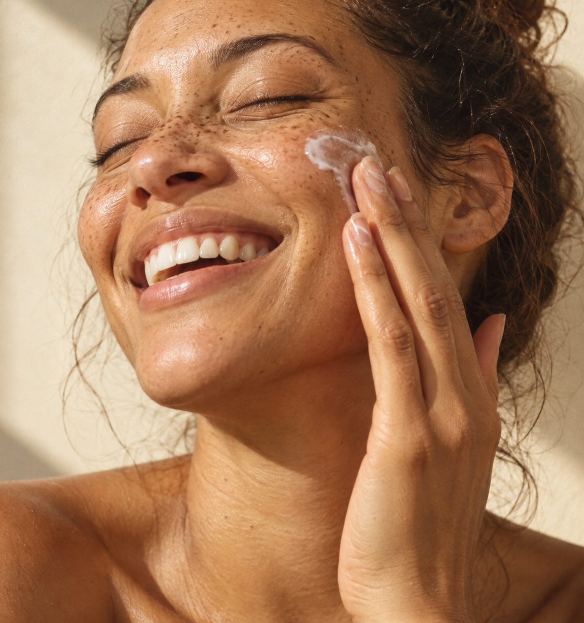 A woman laughing as she smooths cream into her cheek in warm morning light